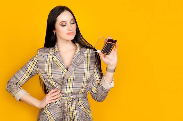 A woman holds a package of artificial lashes, a master of eyelash extension on a bright yellow wall, with copy space. The concept of eyelash extension.