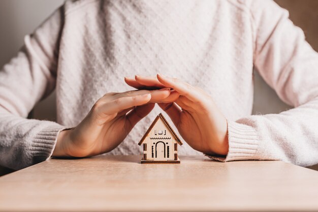 女性は明るいピンクの背景に太陽と彼女の手で木造住宅を保持し、保護します。甘い家