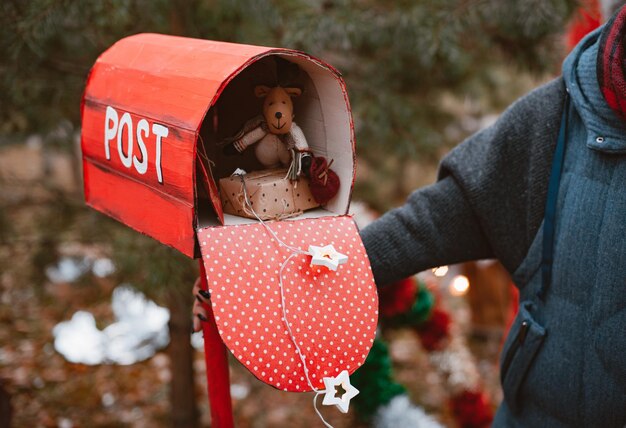 Woman holds a red polka dot retro mailbox with Christmas gifts toys as well as a congratulatory letter from Santa on a background of fir trees.