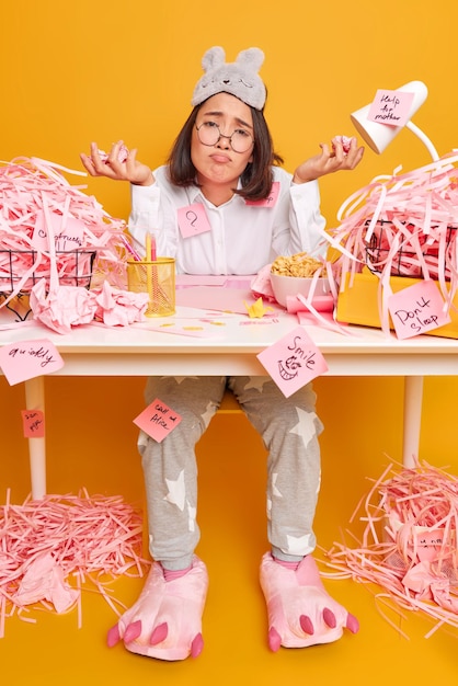 woman holds rumpled paper sits at messy desktop faces problem while doing creative task wears domestic clothes isolated on yellow has much work.