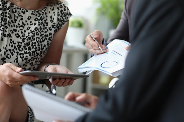 Woman holds tablet in hands next to men with ballpoint pen and documents in hands in office closeup