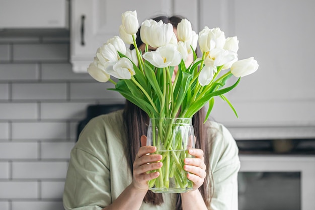 A woman holds a vase with white tulips in a kitchen interior