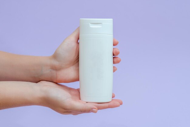 Woman holds a white bottle cream lotion gel shampoo for cosmetic products isolated on lilac background