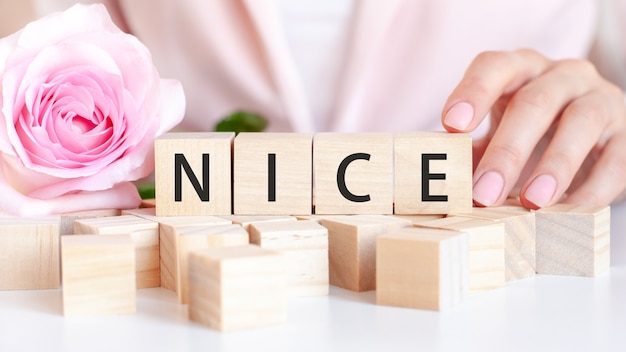 A woman holds a wooden cube with the text of Nice in her hand. On the wooden cubes there is a living rose flower. Pink table, front view. Business and economic concept