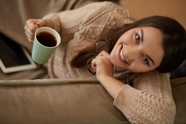 녹색 컵에서 커피를 마시는 태블릿을 사용하여 소파 근처 카펫에 앉아 집 옷을 입은 여성