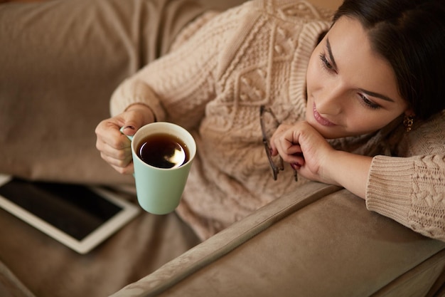 Woman in home clothes sitting on the carpet near the sofa using a tablet drinking coffee from a green cup