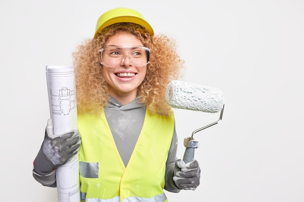 woman house painter holds paint roller and blueprint busy making repairing dressed in working clothes wears protective glasses helmet gloves