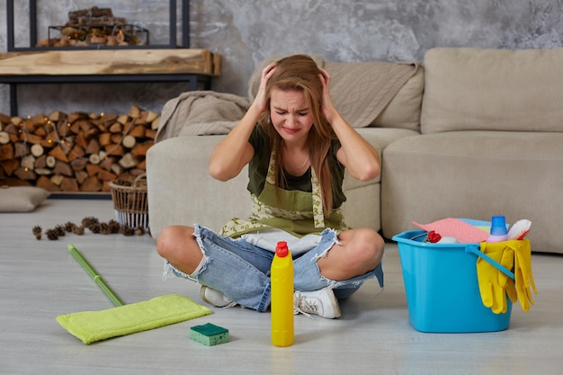 Женщина-домохозяйка чувствует себя уставшей, сидя на полу в гостиной после уборки дома синим ведром, наполненным химикатами и средствами для уборки