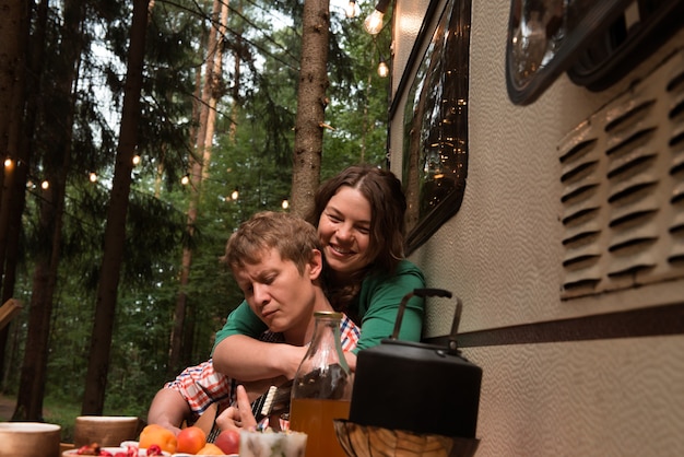 Woman hugging man playing guitar close to trailer in forest summer camping