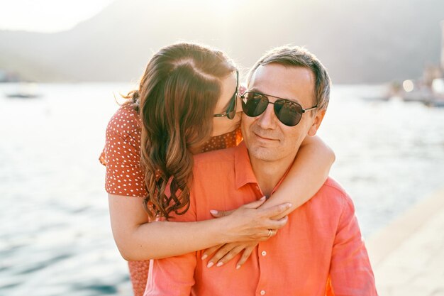 Photo woman hugs the shoulders of a man from behind and kisses his cheek sitting on the pier