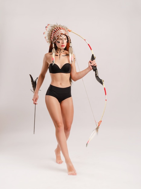 Женщина в образе коренных народов Америки с луком и стрелами позирует на белом фоне