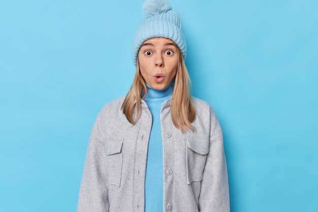 woman impressed by big announcement stares in awewears knitted hat and grey jacket isolated on blue. onwhelmed female model feels concerned.