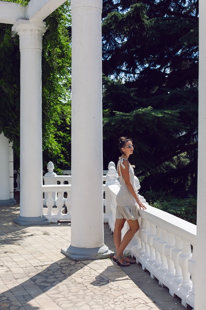 Фото Женщина в коротком платье и солнечных очках стоит у забора и белой колонны на природе в крыму летом