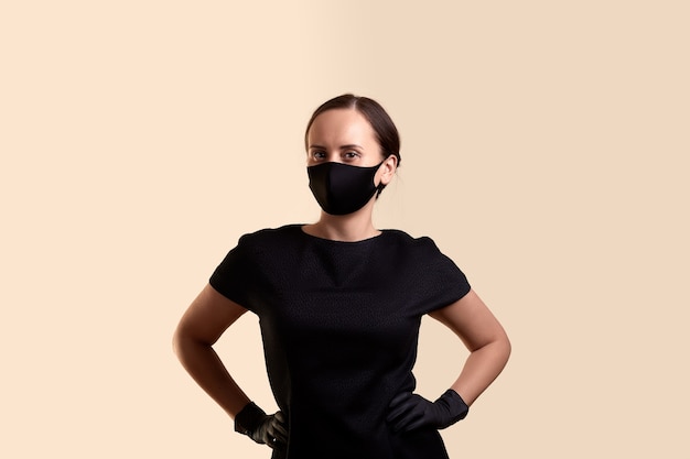 写真 黒のドレスのフェイスマスクとラテックス手袋を着用した女性は、腕を腰に当ててベージュの壁の上に立っていることを誇示します