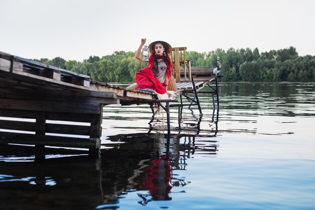 写真 アジア風の服と帽子をかぶった女性がランタンを持って川の木の橋の上に座っている