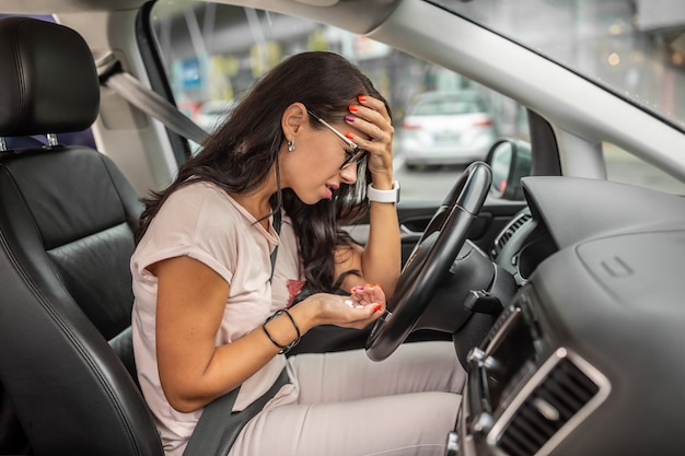 Женщина в отчаянии держит голову, сидя на водительском сиденье с горсткой таблеток в другой руке.