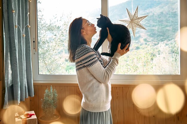 写真 クリスマスを祝う準備をしている間に窓の近くで黒い猫を抱きしめる祭りの気分の女性