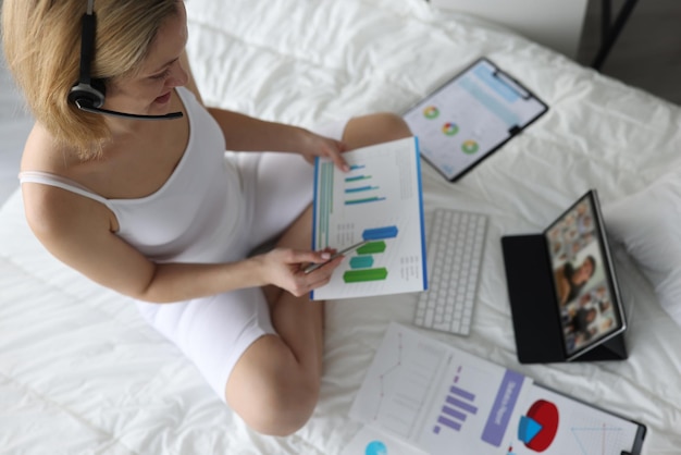 Фото Женщина в наушниках держит графики, сидит на кровати и проводит онлайн-конференцию