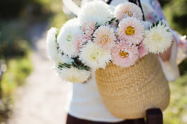 写真 ウクライナの伝統的なシャツを着た女性は、わらのバッグに白とピンクの菊の花束を保持します秋のコンセプト花と日光の下でフィールドを歩く