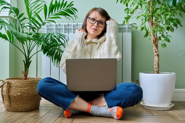 Фото Женщина в теплом свитере с помощью ноутбука сидит рядом с отопительным радиатором дома