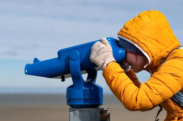 写真 コイン式の双眼鏡を使って海を眺める黄色いコートを着た女性