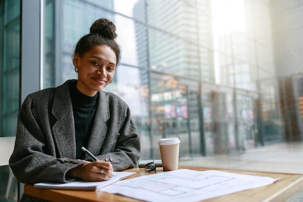Woman interior designer making notes while sitting in cozy cafe near window and looking at camera