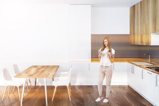 Woman in interior of kitchen with white and gray walls, wooden floor, white countertops with built in sink and cooker and wooden cupboards. Wooden table with white chairs. Toned image