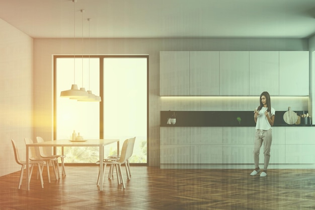 Foto donna all'interno della cucina moderna con pareti bianche, pavimento in legno, controsoffitti e armadi bianchi e tavolo da pranzo bianco con sedie. immagine tonica doppia esposizione