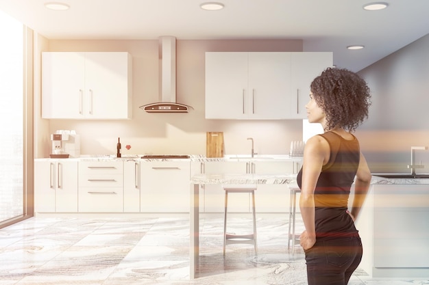 Woman in interior of stylish kitchen with beige walls, white marble floor, large windows, white countertops with built in sink and oven, coffee machine and white bar with stools. Toned image