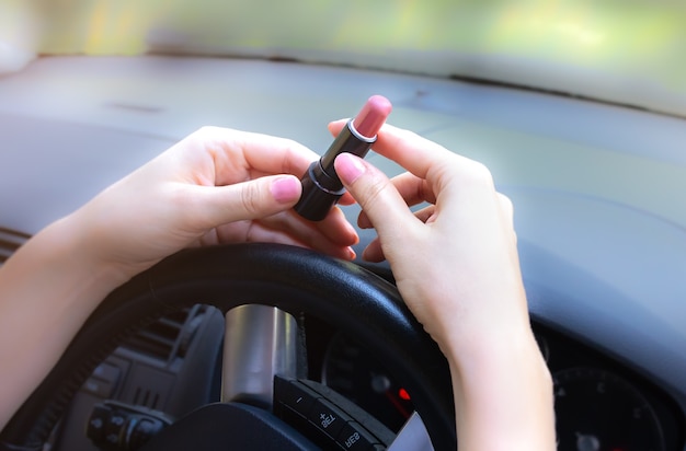 女性が車の中で化粧をしています。ホイールの後ろに口紅を持つ少女。道路上の危険な状況。事故の可能性。