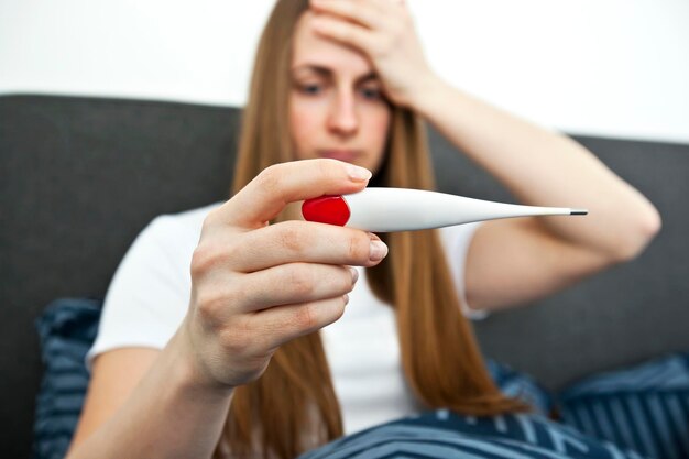 The woman is checking her temperature lying in the bed looking on thermometer