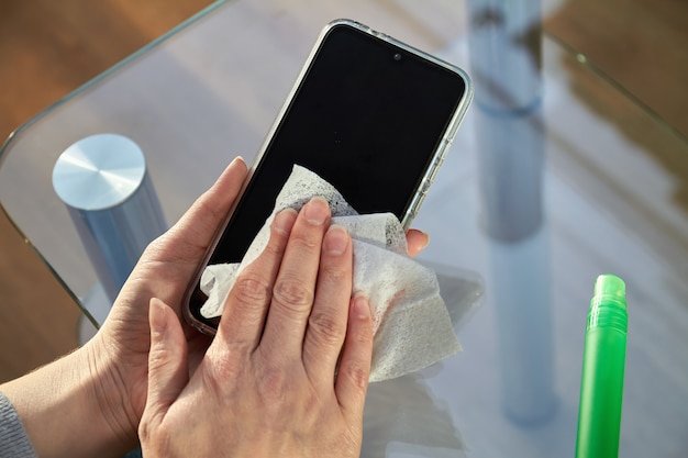 La donna sta pulendo uno smartphone con un getto di disinfettante per mani aerosol di alcol e un panno usa e getta a casa. precauzioni riguardanti i virus