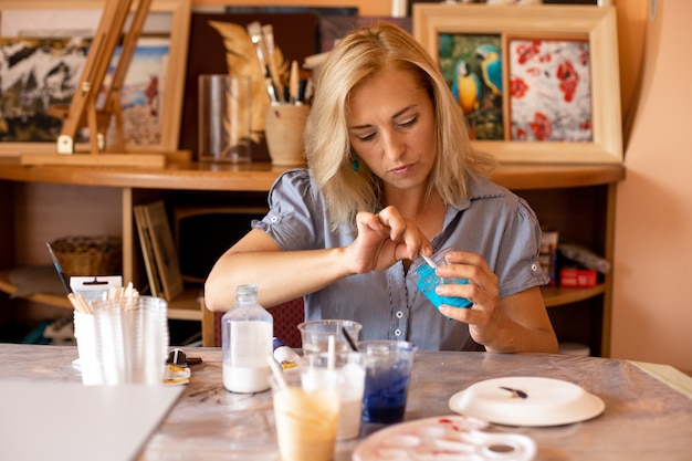 Woman is concentrating on stirring turquoise paint with cup and preparing for work. Work in creative workshop. Interior paintings. Design and inspiration. Work at home. Lifestyle