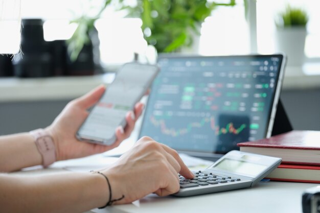 Woman is counting on calculator and holding mobile phone with charts of cryptocurrency rate