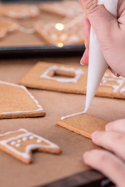 Woman is decorating gingerbread cookies house with white frosting icing cream topping on wooden table background baking paper in kitchen close up macro