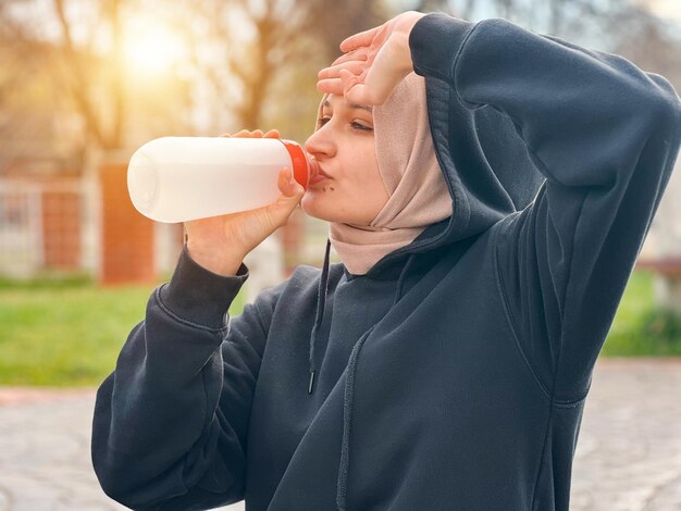 A woman is drinking water from a bottle and wearing a hijab.