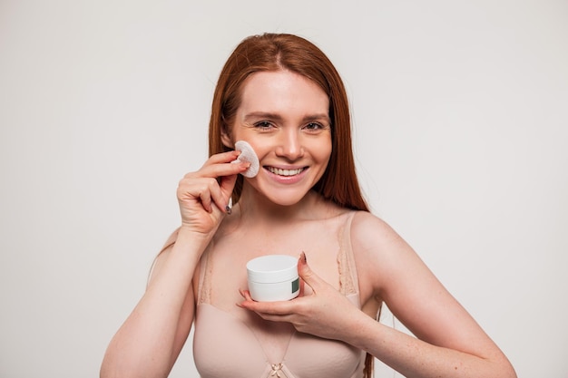 A woman is holding a cream bottle and brushing her face.