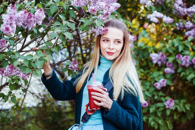 Woman is holding glass of vitamin smoothie in her hands by a floral tree