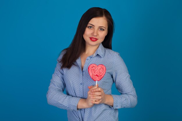 Woman is holding a pink heart-shaped lollipop near her heart aganist of blue background