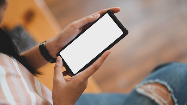 A woman is holding a white blank screen smartphone while sitting in the sitting room