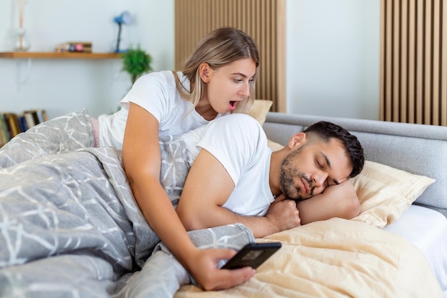 Woman is jealous and suspicious and spies in her partner's smartphone whiles he's sleeping in bedroom The wife is spying on her husband's phone while he sleeps The concept of distrust jealousy