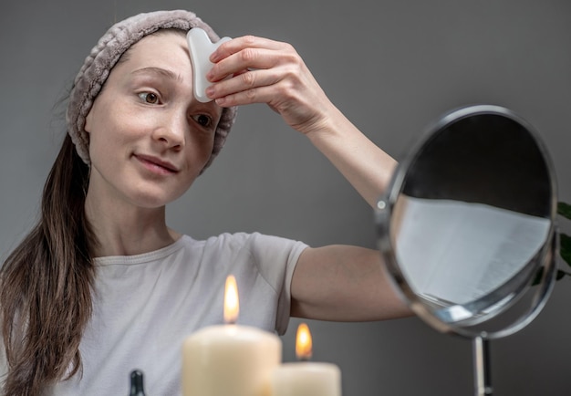 Woman is looking in the mirror and doing a massage with a gouache scraper massager with oil There are burning candles on the table Concept of facial care aromatherapy morning and evening rituals