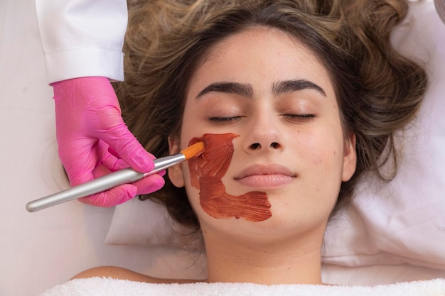 Woman is lying in the cosmetologist s office on the procedure of moisturizing her face The cosmetologist applies a moisturizing mask to the patient's face