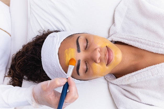 Woman is lying in the cosmetologist s office on the procedure of moisturizing her face. The cosmetologist applies a moisturizing mask to the patient's face.