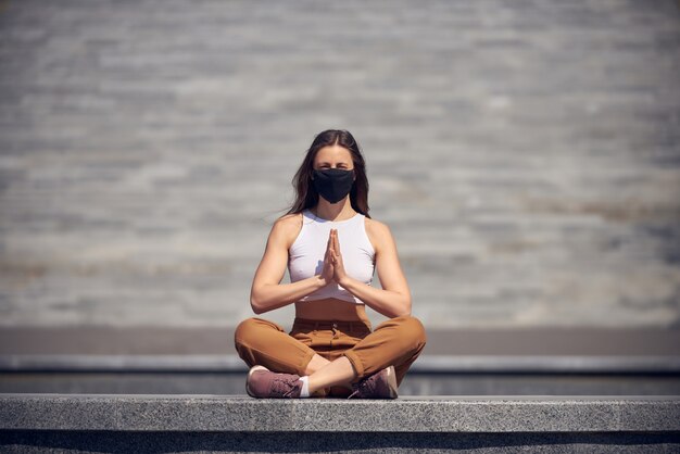 Woman is meditating at empty public place in a black mask. Epidemic of Coronavirus concept photo