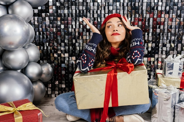 The woman is perplexed by the presented gift Beautiful brunette in a Christmas hat scarf and sweater on a silver background