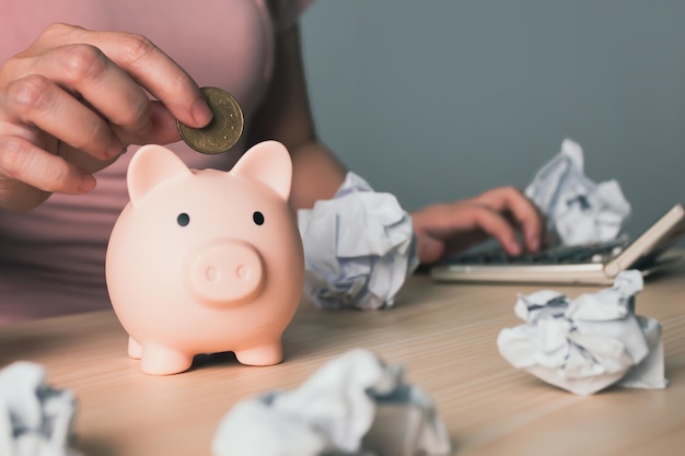 Woman is putting money or coin into piggy bank and using calculator to calculate amount of money for saving plan with crumpled paper Saving money investment banking and finance concept