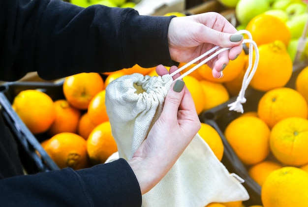 女性は再利用可能な買い物袋にオレンジを入れています。ゼロウェイスト。生態学的および環境に優しいパケット。キャンバスとリネンの生地。自然の概念を保存します。スーパーマーケットでのプラスチックの使い捨てはありません。