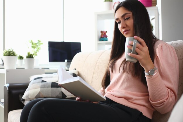 Woman is reading book and drinking from cup sitting on couch