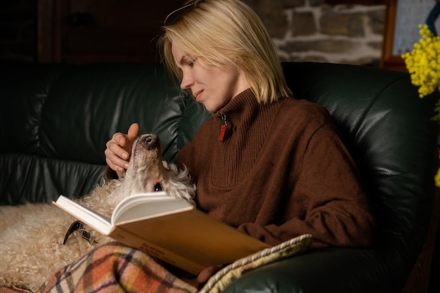 A woman is reading a book on the sofa There is a dog nearby Education reading concept Home cozy interior Attractive woman in her forties